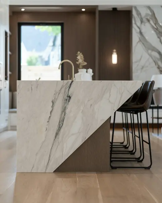 A kitchen island faced in veined white stone with dark leather stools.