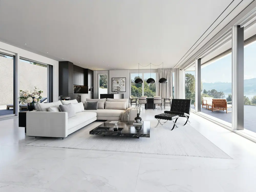 A pale living room with a long white sofa and glass walls open to a lake and hills.