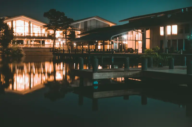 A lakefront house with tall glass gables lit from inside at dusk, its dock and reflection in still water below.