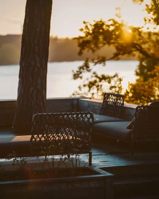 Outdoor chairs on a lakeside terrace at sunset, the water bright behind a tree.