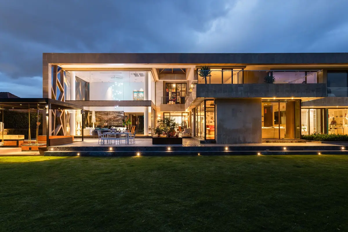 A two-storey glass and concrete house lit from inside at dusk, seen across its lawn.